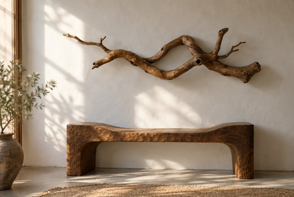 A twisted branch solid wood wall sculpture hanging above a carved wood bench on a white adobe wall, lit by afternoon sun